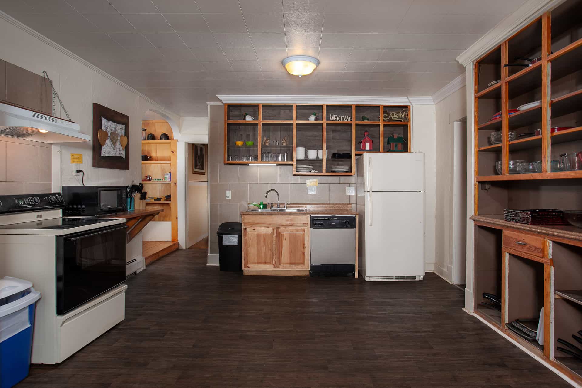 A functional kitchen with white appliances, wooden cabinetry, and open shelving, connected to a dining area and another room in a lodge setting.