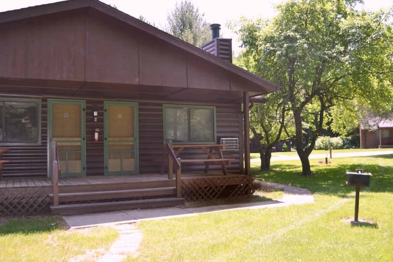 A rustic brown cabin with a front porch and green doors, nestled in a grassy area with trees, featuring a grill and a paved walkway.
