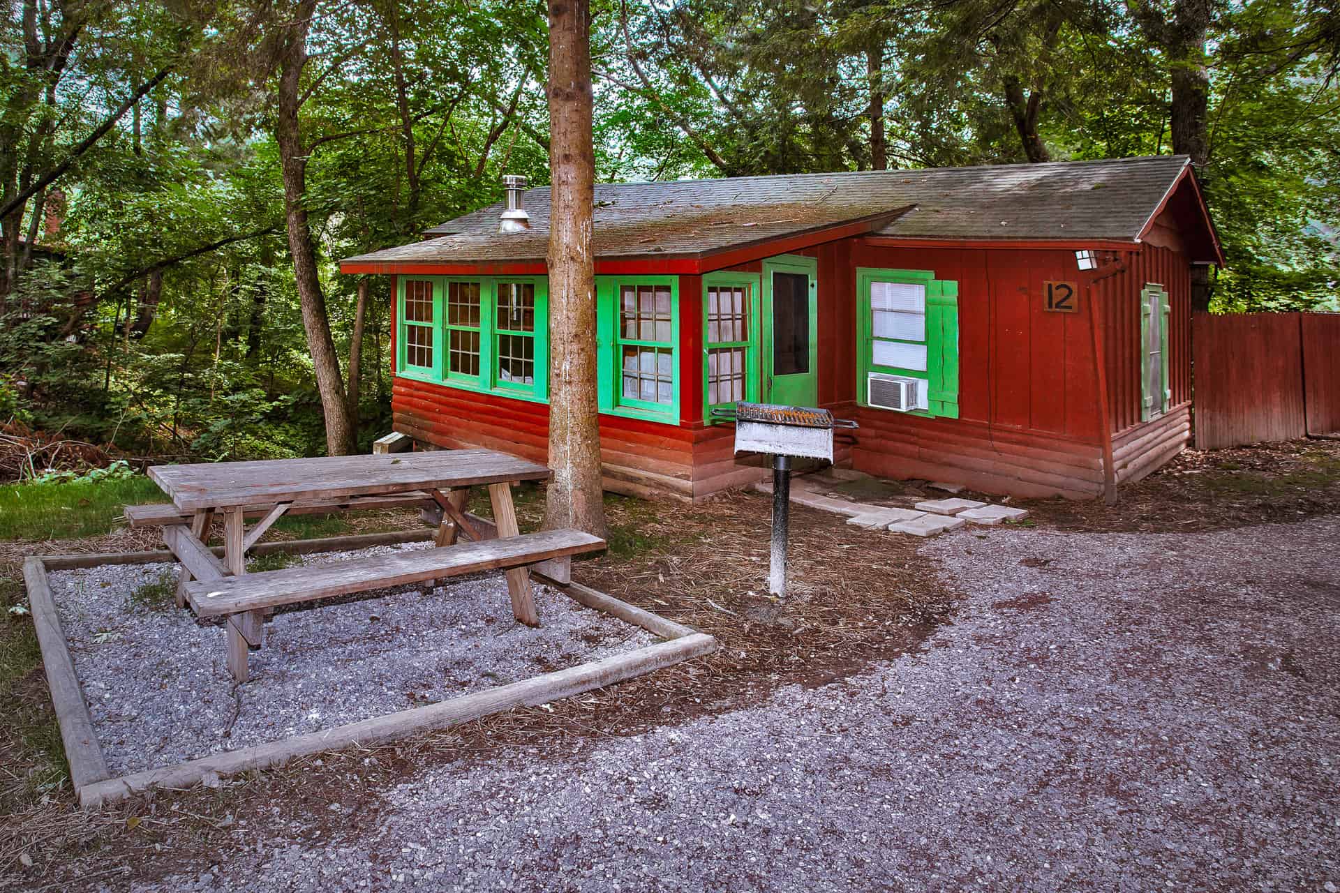 A quaint red cabin with vibrant green trim and shutters, nestled amongst trees, featuring a picnic table and grill outside.