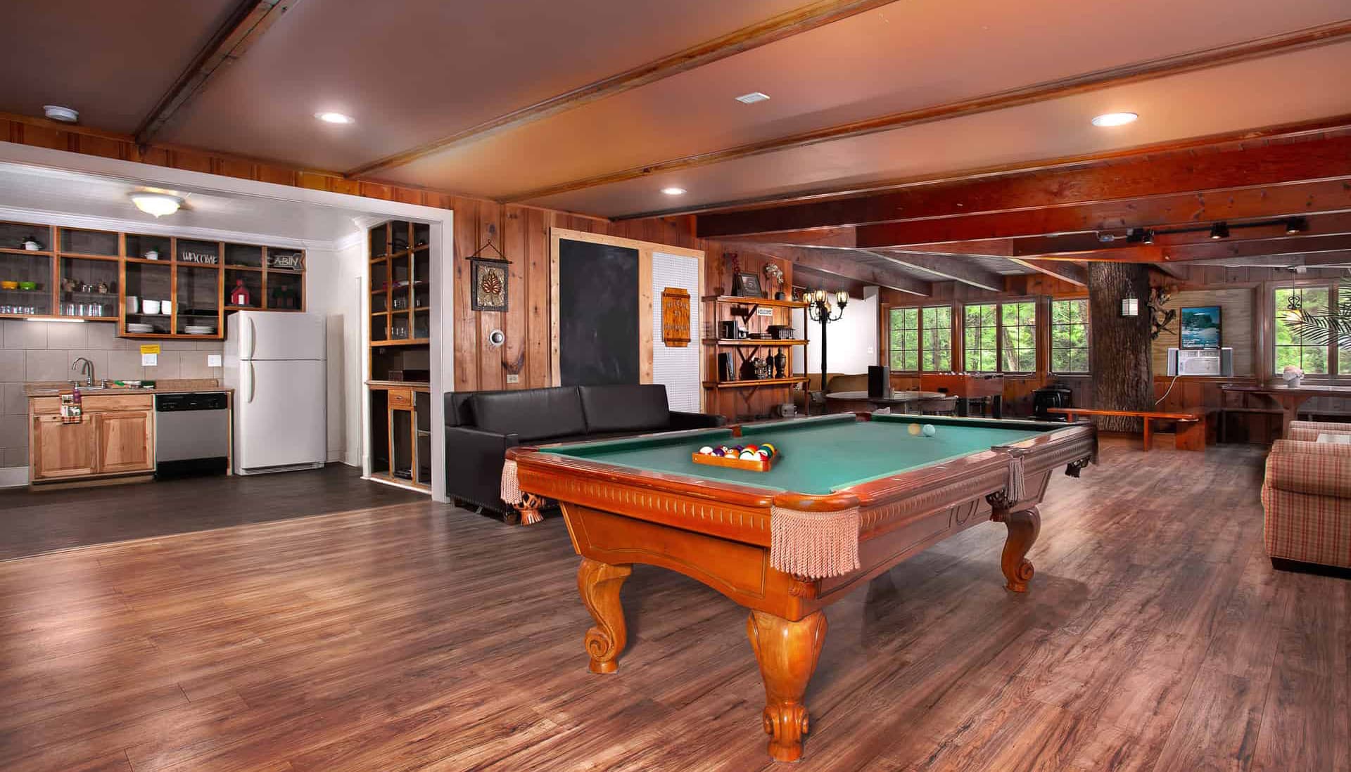 Interior of a cozy cabin featuring a pool table, sofa, kitchenette, and large windows.
