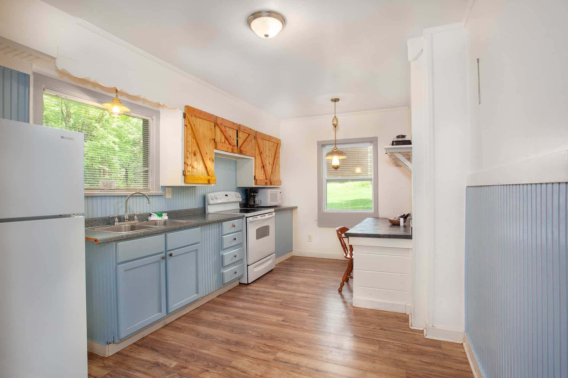 A quaint kitchen with light blue lower cabinets, a white refrigerator and stove, and a small dining nook, offering a bright and airy feel.