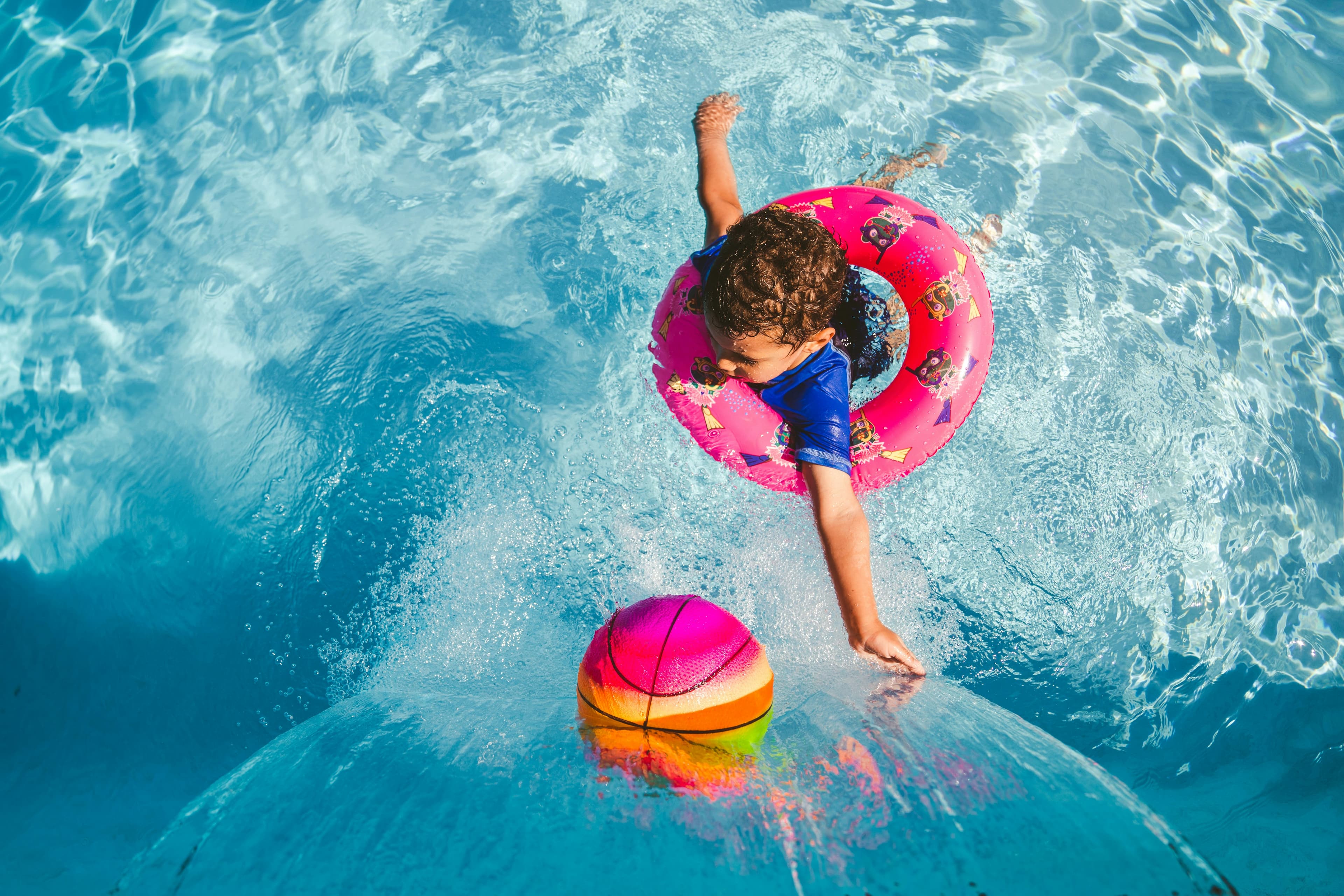 A child in a pink float splashes into a swimming pool while reaching for a colorful beach ball.