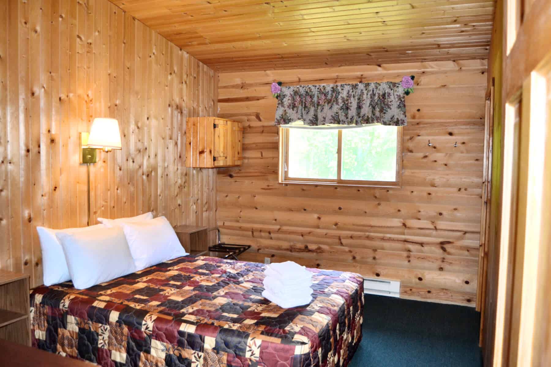 A cozy cabin bedroom featuring knotty pine walls, a bed with a colorful quilt, a small window with a valance, and a wall-mounted lamp.