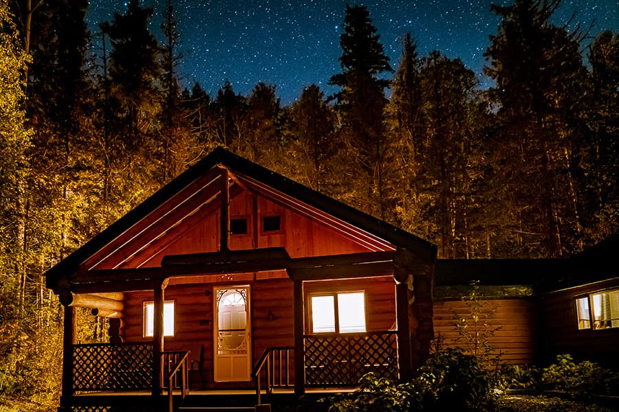 A warmly lit log cabin surrounded by trees under a starry night sky.
